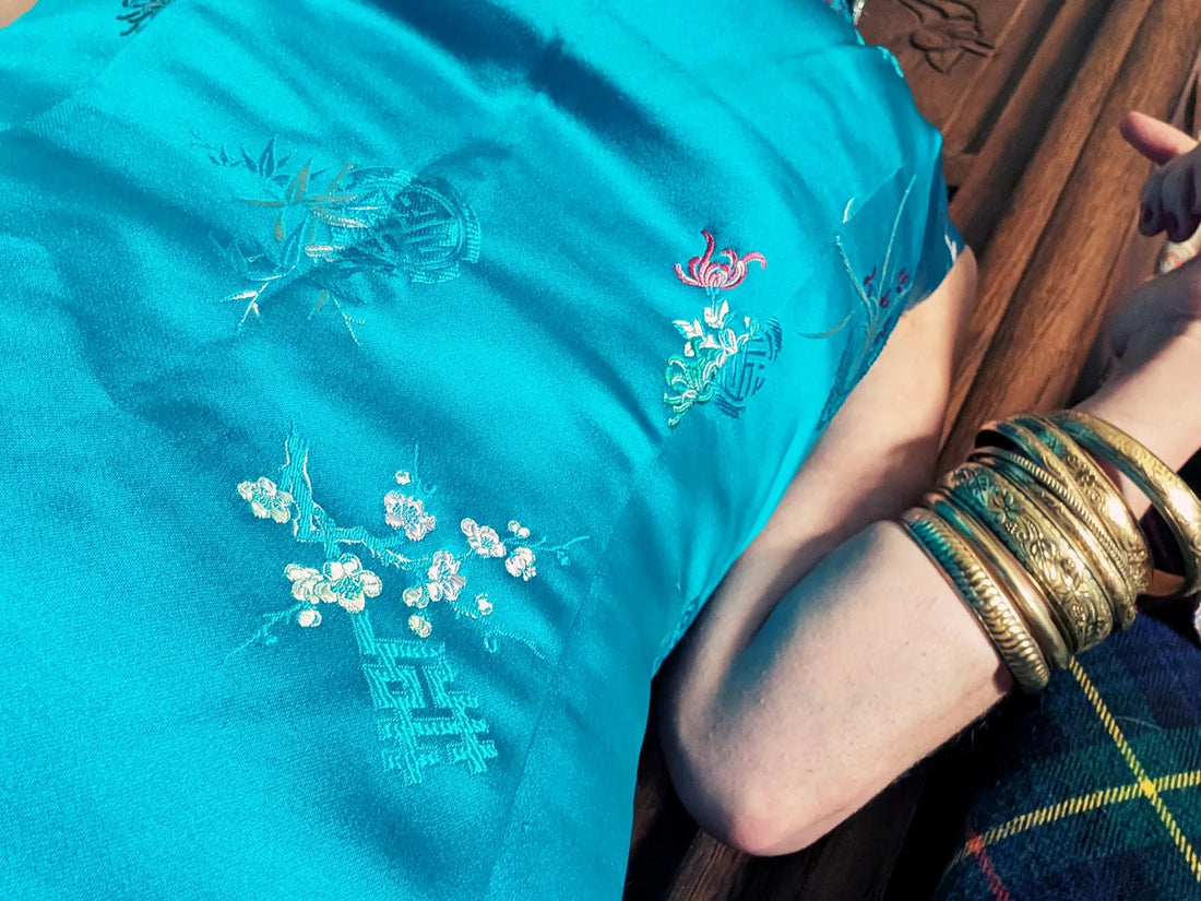 Tissu de robe qipao vintage en satin turquoise, brodé de motifs floraux délicats, reposant sur un bras orné de bracelets dorés