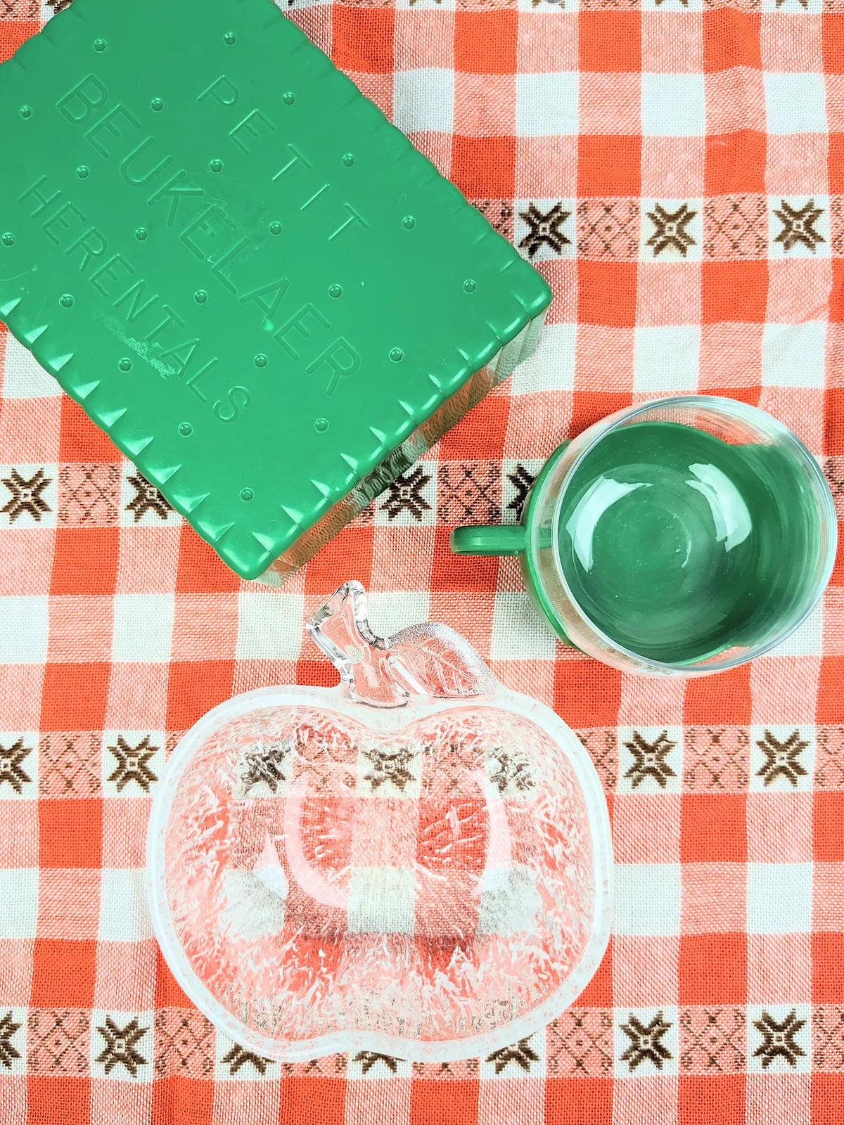 Table avec nappe à carreaux rouge  orange vintage avec boîte à tartine verte plastique, plat en forme de pomme en verre et tasse en verre et plastique vert