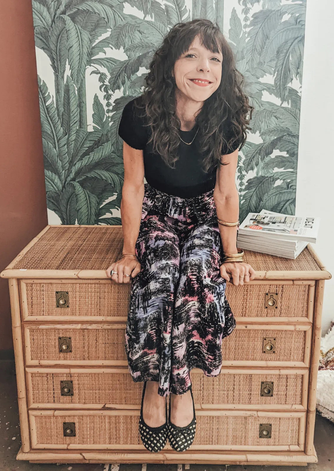 Stéphanie Meyer souriante assise sur une commode en rotin, devant un papier peint végétal, portrait Les Passantes Vintage
